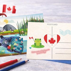 Puede incluir: Dos postales con temas canadienses. Una presenta una rana de dibujos animados con una bandera canadiense, la otra un oso de dibujos animados en un lago. Una taza roja de café, lápices rojos y azules y banderas canadienses también están en la imagen.