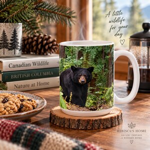 May include: A white ceramic mug featuring a photographic print of a black bear standing in a lush green forest. The mug has a smooth handle and sits on a round wooden slice coaster. The text A little wildlife for your day is printed above.