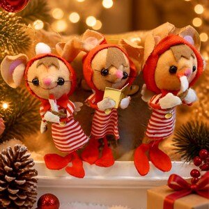 May include: Three plush mouse ornaments wearing red hooded outfits with white striped shirts and red shoes. One mouse holds a white candle, another holds a small gold book, and the third holds a white object. They have pink noses and large ears.