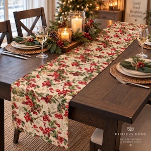 May include: A rectangular table runner featuring a festive pattern of red holly berries, green leaves, and small white snowflakes on a cream-colored fabric. The runner is centered on a dark wood table with place settings and a centerpiece.