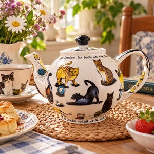 Peut inclure: Une théière en céramique blanche ornée d'illustrations colorées de divers chats dans différentes positions, dont un tabby roux et un chat noir. La théière possède un bouton de couvercle noir et une inscription sur le bord : "I went to London to look at the Queen."