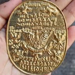 Pode incluir: Um pingente dourado em forma oval com detalhes intrincados em relevo. O pingente apresenta texto e um desenho central de um pássaro com as asas abertas. Uma pequena argola na parte superior para fixação. O pingente é segurado em uma mão.
