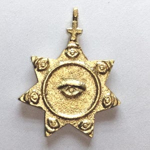 Pode incluir: Pingente dourado com design de estrela. O pingente apresenta um círculo central com um símbolo de olho e é rodeado por uma estrela de sete pontas. O pingente tem uma argola com uma cruz no topo.