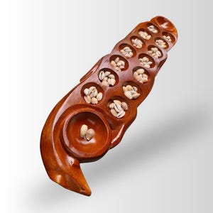 May include: A polished brown wooden mancala game board featuring two parallel rows of six circular pits and two larger end storage bowls. The board is filled with small, off-white cowrie shells distributed throughout the various carved indentations.