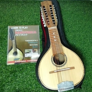 Könnte beinhalten: Eine hellfarbene hölzerne Bandurria mit zwölf Saiten, einem runden Schallloch und einem Metallsaitenhalter, begleitet von einem spiralgebundenen Lehrbuch mit dem Titel Learn to Play Small Bandurria Piccollo with Fundamentals Techniques Notations Scales.