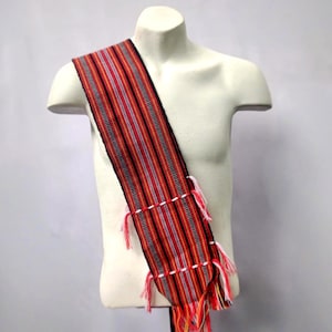 Può includere: Una fascia tessuta con un motivo a righe rosse, nere e bianche, esposta su un manichino bianco. La fascia ha nappe decorative rosse, gialle e bianche nella parte inferiore. Il tessuto è probabilmente fatto a mano.