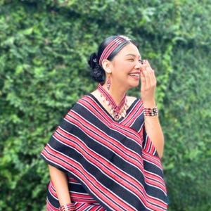 May include: A woman wearing a traditional outfit with a red, black, and white striped shawl. She is also wearing a matching headband, earrings, and bracelets. The woman is smiling and laughing.