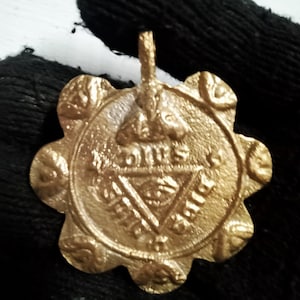 Könnte beinhalten: Ein goldfarbener Metallanhänger mit einem runden Design, das eine Krone, ein Dreieck mit einem Auge darin und die Worte "IN HOC SIGNO VINCES" zeigt. Der Anhänger hat einen gewellten Rand.