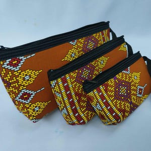 Peut inclure: Ensemble de trois pochettes zippées de tailles différentes. Les pochettes sont en tissu marron avec un motif géométrique coloré jaune, rouge et blanc. Chaque pochette a une fermeture éclair noire et une dragonne noire.