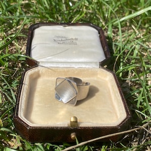 May include: A silver-toned ring featuring a square-shaped top with a brushed metal finish, resting inside an open, dark brown, octagonal jewellery box with a cream-coloured velvet interior lining and visible text printed on the inner lid.