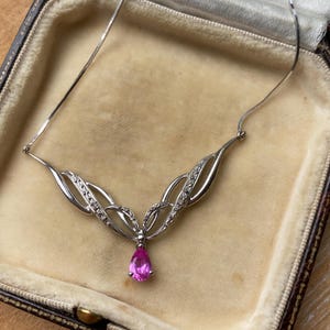May include: A silver necklace with a delicate design, featuring a teardrop-shaped pink gemstone pendant. The necklace is displayed in a vintage jewellery box with a beige interior.