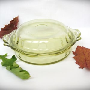 Puede incluir: Una fuente de vidrio verde claro con tapa a juego. La fuente tiene dos asas y un borde ligeramente festoneado. La tapa tiene un pequeño pomo. Hojas de otoño rodean la fuente.