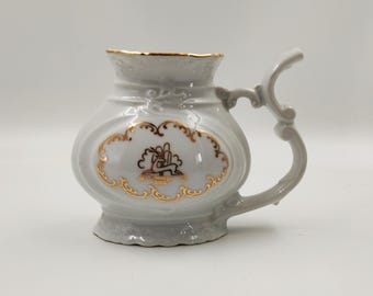 Puede incluir: Una taza de cerámica blanca con borde y asa dorados decorativos. La taza presenta un emblema dorado de un ciervo y detalles dorados ornamentados. La taza tiene una forma única y probablemente sea para uso decorativo.