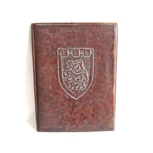 Op de afbeelding: Een bruin lederen gebonden boek met een reliëfdesign van een schild met drie leeuwen. Het schild is omgeven door een decoratieve rand.