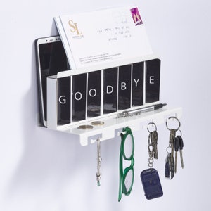 May include: White wall-mounted key holder with a shelf and hooks. The shelf has a compartment that says "GOODBYE" and a phone holder. There are keys, glasses, and coins on the hooks and shelf.