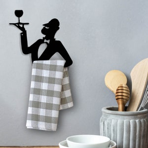 May include: Black metal silhouette of a waiter holding a tray with a wine glass, used as a towel rack. A gray and white checkered kitchen towel is hanging from the rack.