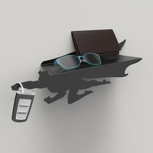 Op de afbeelding: Een zwart metalen plank met superheldenthema en een cape-ontwerp. De plank bevat een bruine portemonnee, blauwe zonnebril en een autosleutel. Het silhouet van de superheld staat in een vliegende pose.