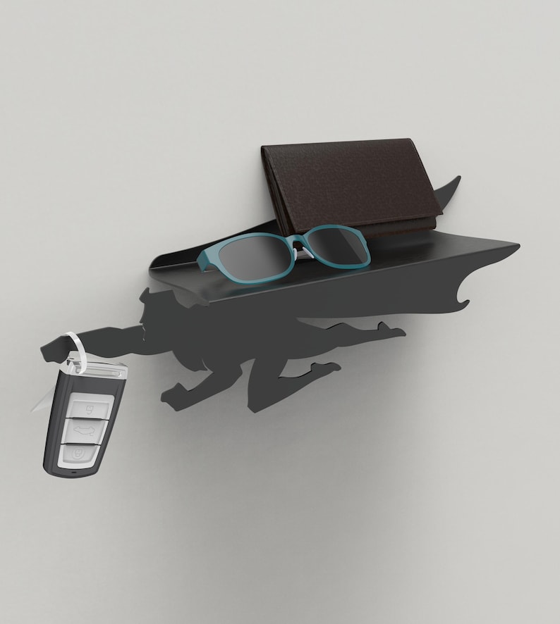 Op de afbeelding: Een zwart metalen plank met superheldenthema en een cape-ontwerp. De plank bevat een bruine portemonnee, blauwe zonnebril en een autosleutel. Het silhouet van de superheld staat in een vliegende pose.