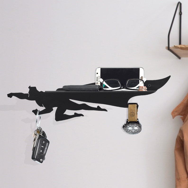 Op de afbeelding: Zwart metalen wandrek in de vorm van een superheld, met een telefoon, bril, portemonnee, horloge en sleutels. Het rek is aan een witte muur bevestigd en biedt een stijlvolle opbergoplossing.