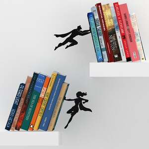 May include: Black silhouette of a superhero holding up a shelf of books. The books have titles like "The Arabian Nights", "The Great Gatsby", "Urban Theory", and "Rebel Thirds".