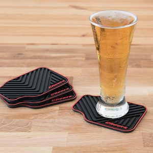 May include: Four black and red silicone coasters with the word "Cheers" printed on each. A glass of beer is sitting on one of the coasters.