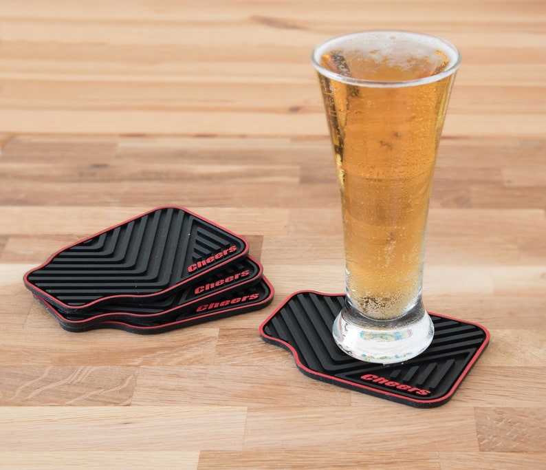 May include: Four black and red silicone coasters with the word "Cheers" printed on each. A glass of beer is sitting on one of the coasters.