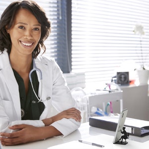 Peut inclure: Une femme portant une blouse blanche avec un st&eacute;thoscope autour du cou est assise &agrave; un bureau. Elle sourit et regarde la cam&eacute;ra. Il y a un support de t&eacute;l&eacute;phone noir et blanc sur le bureau avec une silhouette d'une personne assise sur une chaise.