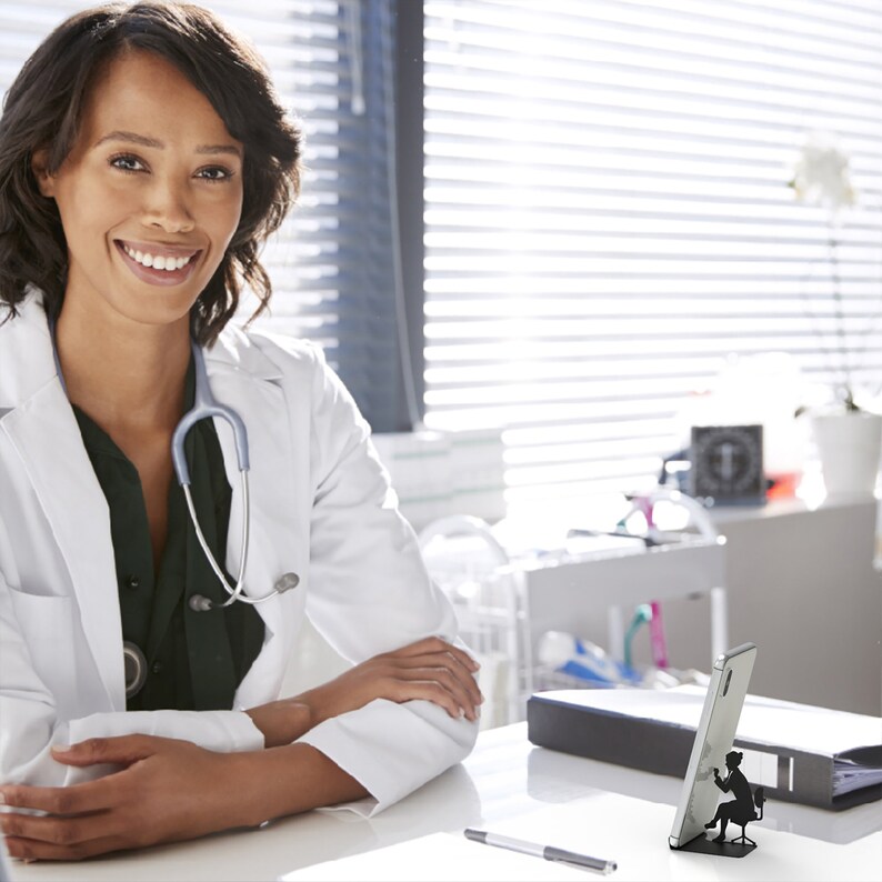 Peut inclure: Une femme portant une blouse blanche avec un st&eacute;thoscope autour du cou est assise &agrave; un bureau. Elle sourit et regarde la cam&eacute;ra. Il y a un support de t&eacute;l&eacute;phone noir et blanc sur le bureau avec une silhouette d'une personne assise sur une chaise.