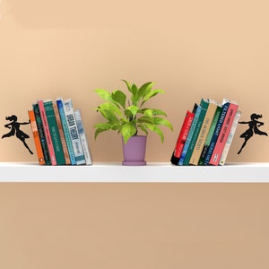 May include: Two black metal bookends shaped like a superhero figure holding books on a white shelf. The bookends are holding a stack of books on each side of a potted plant with green leaves and a purple pot.