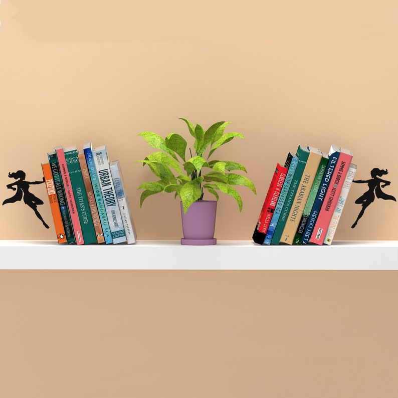 May include: Two black metal bookends shaped like a superhero figure holding books on a white shelf. The bookends are holding a stack of books on each side of a potted plant with green leaves and a purple pot.