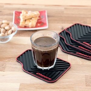 May include: Four black and red square coasters with the word "Cheers" printed on each. A glass of dark coffee sits on one of the coasters.