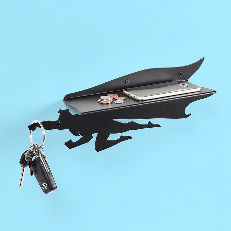 Op de afbeelding: Een zwart metalen wandplank met superheldenthema met een ingebouwd plankje voor een telefoon en munten. De superheld houdt een sleutelring met sleutels vast. De plank is aan een lichtblauwe muur bevestigd.