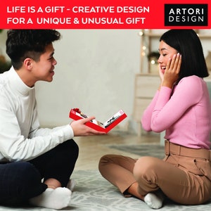 May include: A man is giving a red gift box to a woman. The box is open and shows a picture inside. The text on the box reads "LIFE IS A GIFT - CREATIVE DESIGN FOR A UNIQUE & UNUSUAL GIFT ARTORI DESIGN".