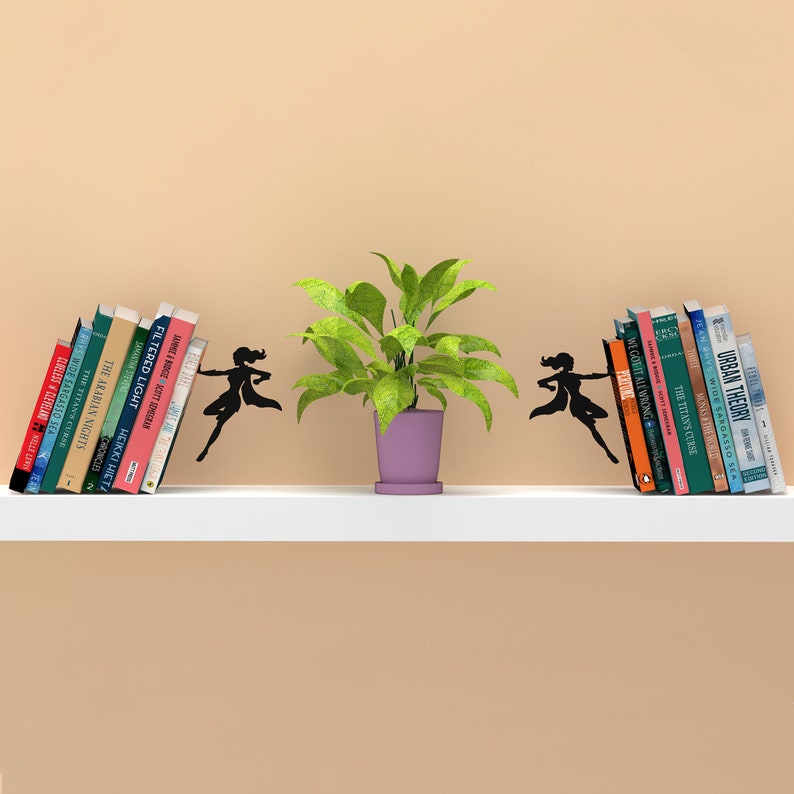 May include: A pair of black metal bookends shaped like women in a superhero pose, holding up a stack of books on a white shelf. A potted green plant sits between the bookends.