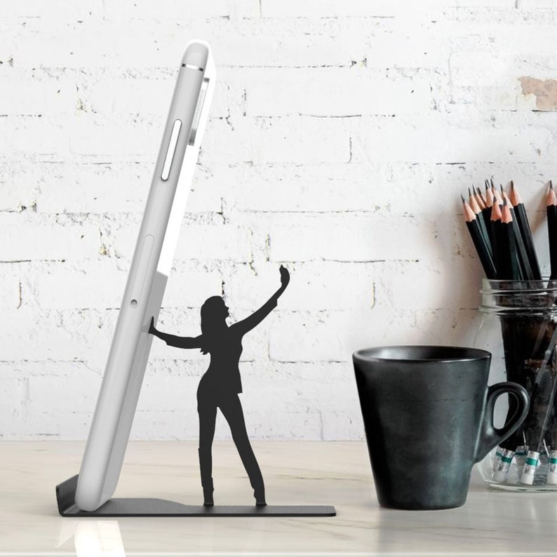 Peut inclure: Support de t&eacute;l&eacute;phone en m&eacute;tal noir en forme de personne prenant un selfie. Le support maintient un smartphone blanc en position verticale.
