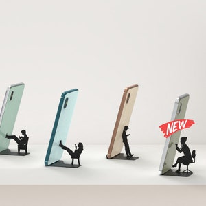 Peut inclure: Six supports de t&eacute;l&eacute;phone de couleurs diff&eacute;rentes avec une silhouette d'une personne tenant un t&eacute;l&eacute;phone ou assise sur une chaise. Les supports sont en m&eacute;tal et ont un design moderne. Le mot "NEW" est affich&eacute; sur deux des supports.
