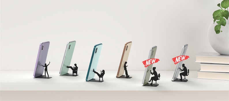 Peut inclure: Six supports de t&eacute;l&eacute;phone de couleurs diff&eacute;rentes avec une silhouette d'une personne tenant un t&eacute;l&eacute;phone ou assise sur une chaise. Les supports sont en m&eacute;tal et ont un design moderne. Le mot "NEW" est affich&eacute; sur deux des supports.