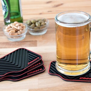 May include: Four black and red silicone coasters with the word "Cheers" printed on each. A glass mug of beer sits on one of the coasters.