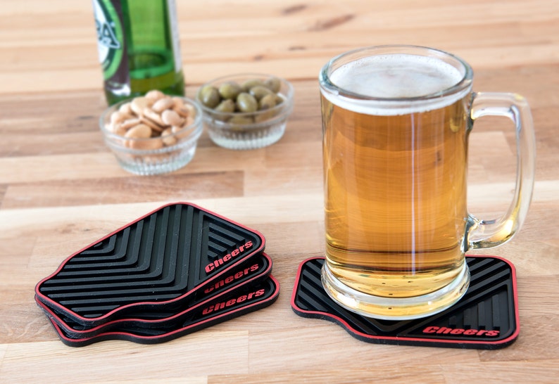 May include: Four black and red silicone coasters with the word "Cheers" printed on each. A glass mug of beer sits on one of the coasters.