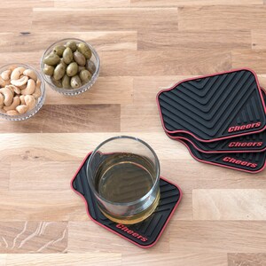 May include: Four black and red coasters with the word "Cheers" printed on each. The coasters are on a wooden surface with two bowls of snacks, one with cashews and one with green olives.