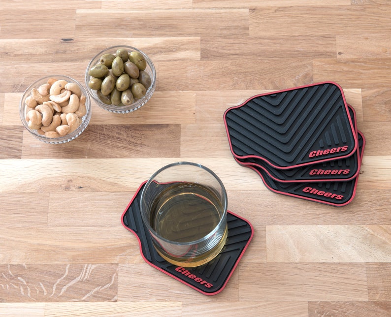 May include: Four black and red coasters with the word "Cheers" printed on each. The coasters are on a wooden surface with two bowls of snacks, one with cashews and one with green olives.