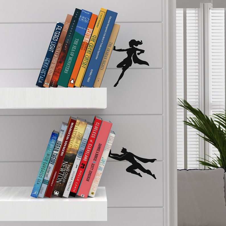 May include: Two white shelves with black metal bookends shaped like superheroes holding up books. The bookends are on a white wall. The books on the top shelf include titles like "The Arabian Nights", "The Great Gatsby", and "The Sea of Monsters". The books on the bottom shelf include titles like "Clueless in Cleveland", "Rebel Thards", and "Manhattan Voyagers".