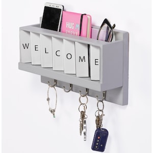 May include: Gray wooden wall mounted key holder with the word "WELCOME" spelled out in white letters. The key holder has five hooks and a shelf for storing small items. There are keys, a phone, and a notebook on the shelf.