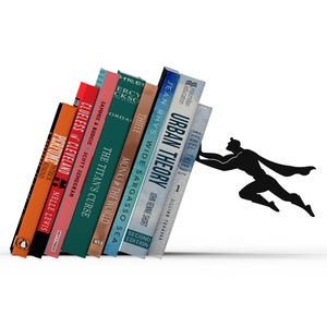 Könnte beinhalten: Ein schwarzer Buchständer aus Metall in Form eines fliegenden Superhelden, der gegen eine Reihe verschiedener Bücher drückt, darunter Titel wie Perfume, Clueless in Cleveland, The Titan's Curse, Monks in the World, Wide Sargasso Sea und Urban Theory.