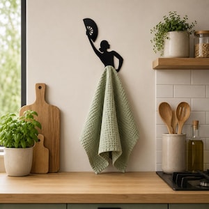 Peut inclure: Un crochet mural en métal noir en forme de silhouette de personne tenant un éventail, avec une serviette vert sauge à texture gaufrée suspendue. Le crochet est fixé sur un mur clair au-dessus d'un plan de travail en bois dans une cuisine.