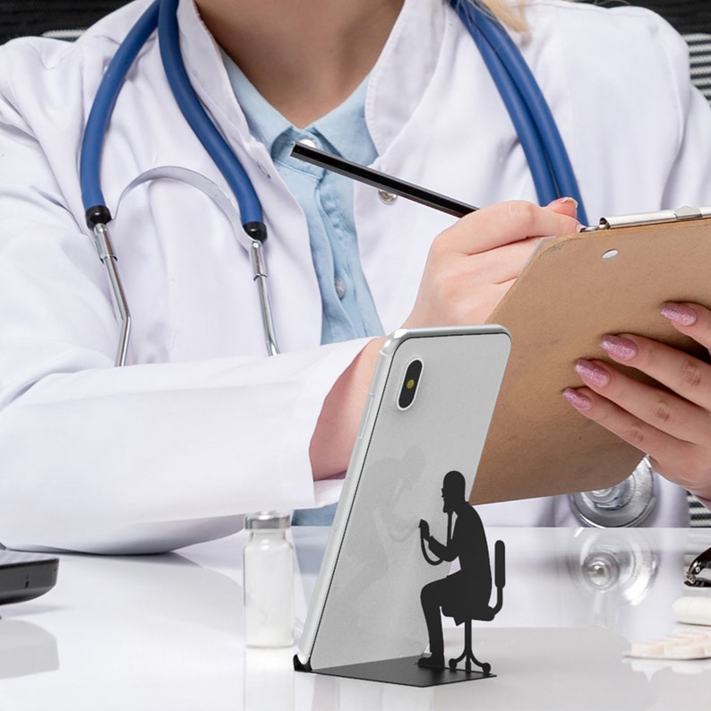 Peut inclure: Un smartphone blanc avec une silhouette noire d'un m&eacute;decin &eacute;coutant la poitrine d'un patient avec un st&eacute;thoscope. Le t&eacute;l&eacute;phone est pos&eacute; sur un support noir.
