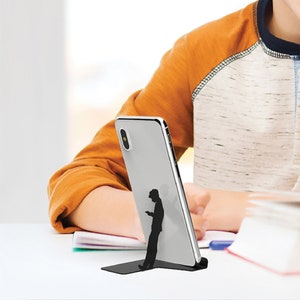 Peut inclure: Un smartphone blanc est pos&eacute; sur un support noir en forme de personne regardant son t&eacute;l&eacute;phone. Le support est sur une table blanche avec un carnet et un stylo.
