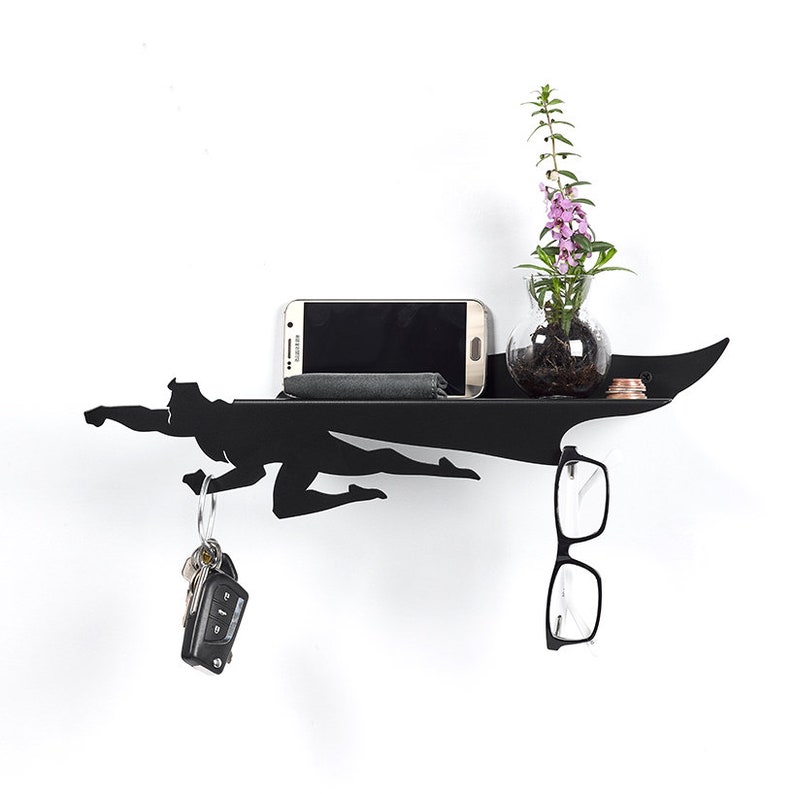 Op de afbeelding: Zwart metalen wandrek in de vorm van een vliegende superheld. Het rek bevat een smartphone, een kleine grijze doek, een kleine glazen vaas met paarse bloemen, sleutels en een bril. Het rek is aan een witte muur bevestigd.