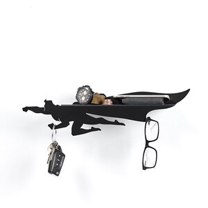 Op de afbeelding: Zwart metalen wandrek in de vorm van een vliegende superheld. Het rek heeft een plank voor een horloge, portemonnee, munten en een telefoon. Sleutels en een bril hangen eronder. Een decoratief en functioneel item.