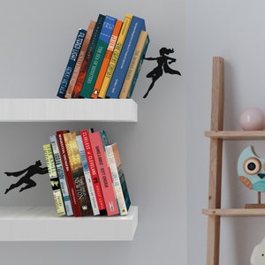 May include: Two white shelves with books on them. The top shelf has a superhero silhouette holding up the books. The bottom shelf has a superhero silhouette holding up the books. The shelves are against a white wall.